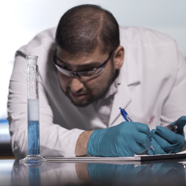 A male scientist in a lab coat watches a graduated cylinder closely