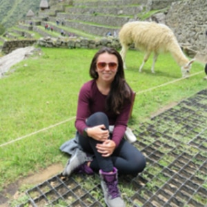 Karen sits on grass near ruins with a llama eating behind her
