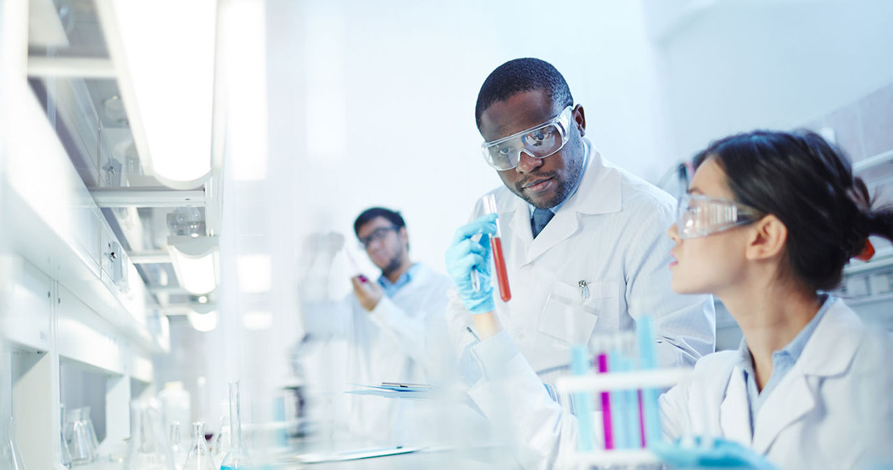 Female Asian laboratory scientist in lab coat and safety goggles showing test tube with red liquid to curious African-American colleague in laboratory. Latin-American scientist in background.Conducting Experiment in Laboratory