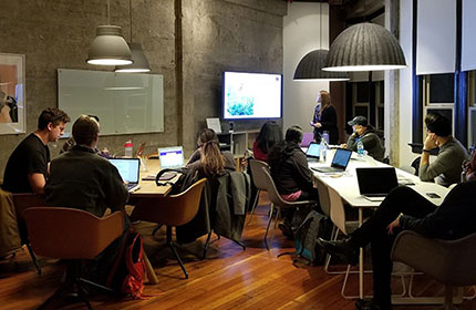 A group of people sit around tables in a modern office, one is presenting from a TV