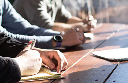 Close up of people sitting around a wooden table while taking notes