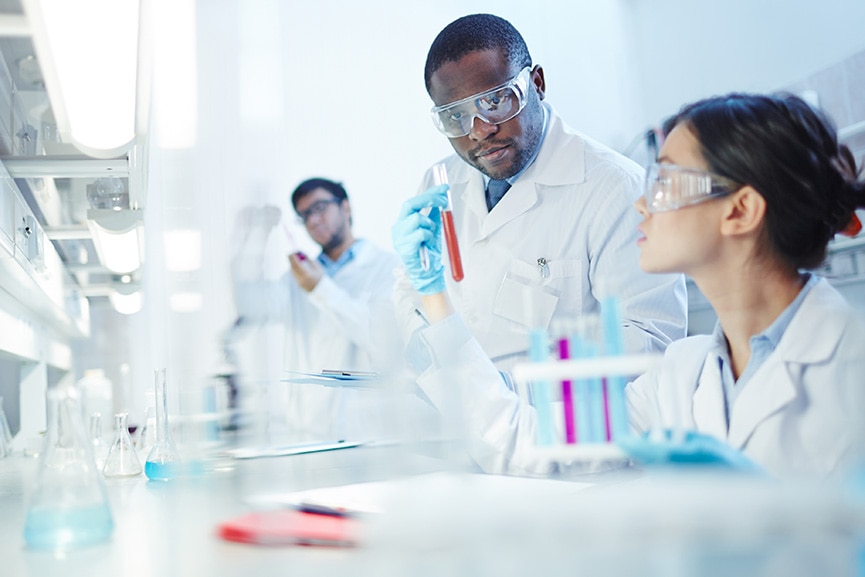 Female Asian laboratory scientist in lab coat and safety goggles showing test tube with red liquid to curious African-American colleague in laboratory. Latin-American scientist in background.
Conducting Experiment in Laboratory