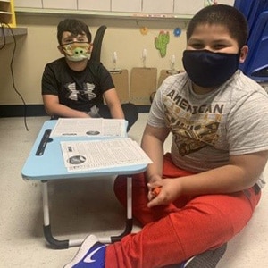 Two students in face masks work together on a lap desk